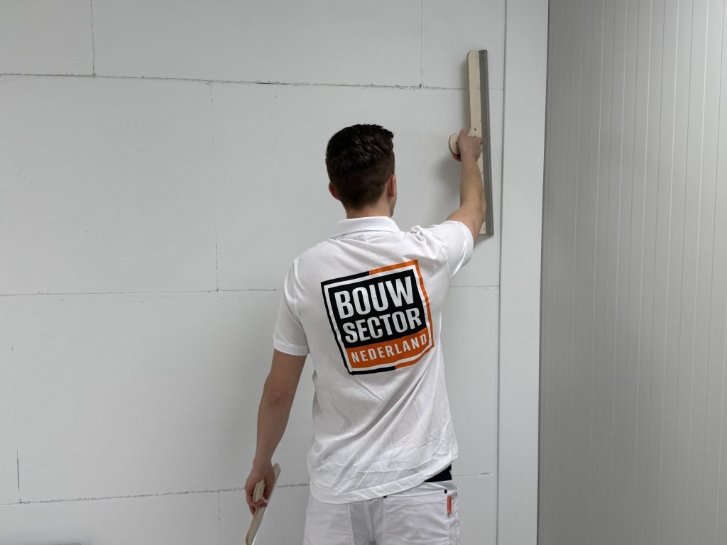 Stukadoor met het logo van Bouwsector Nederland op zijn rug trekt een spaan over een muur van gipsblokken