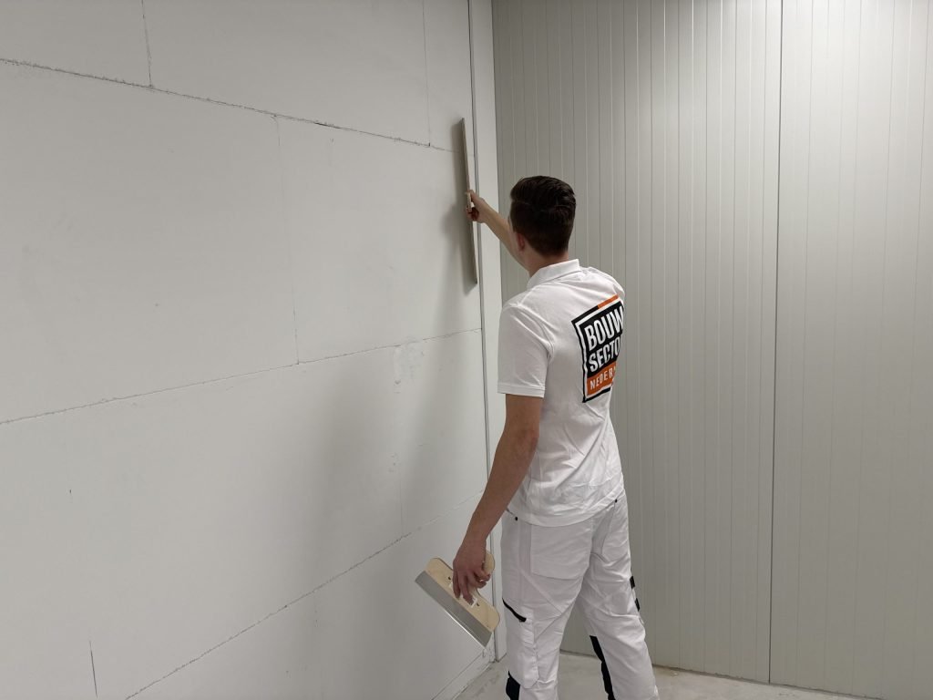 Stukadoor werkt met een lange spaan langs de hoek van een wand, met een plamuurmes in zijn andere hand