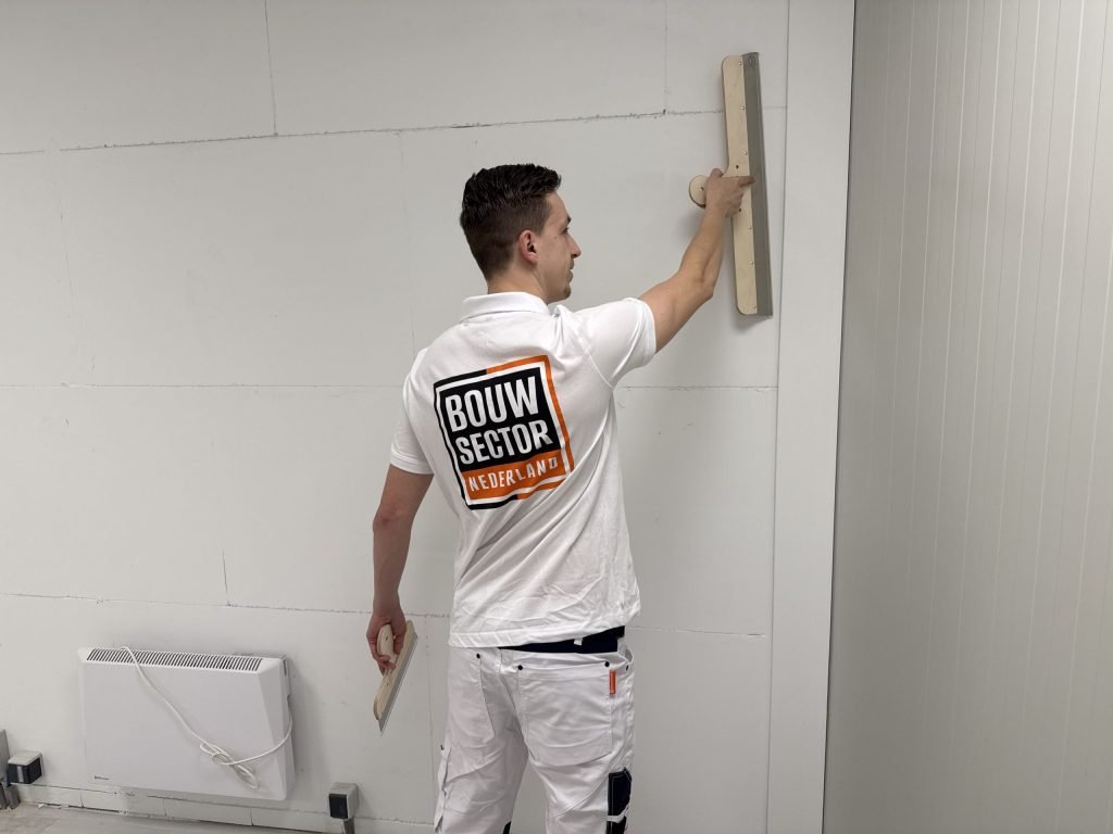 Stukadoor in wit tenue haalt een lange spaan over een wand naast een elektrische kachel